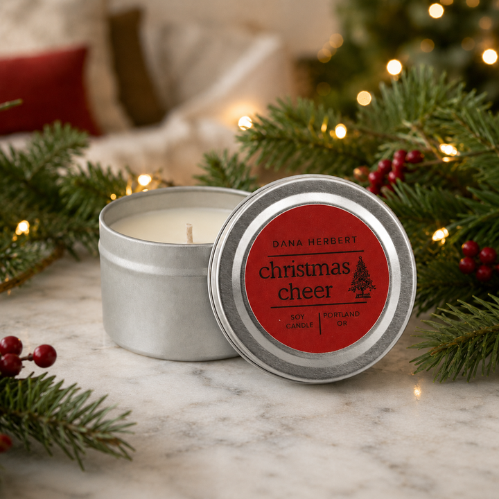 A silver metal tin candle with its lid off, showing a red label that reads 'DANA-HERBERT christmas cheer' and 'poppyseed oil.' The candle is styled on a marble surface with pine branches, red berries, and warm string lights in the background.