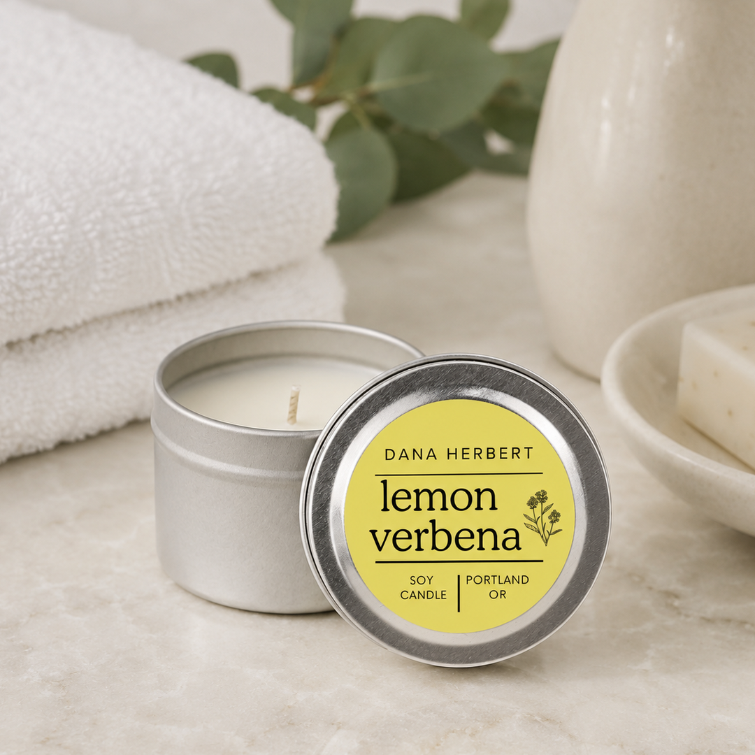 A silver metal tin candle with its lid placed in front, showing a yellow label that reads 'DANA HERBERT lemon verbena SOY CANDLE OR PORTLAND OR.' The candle sits on a light stone surface with a folded white towel, green leaves, and a ceramic vase in the background.