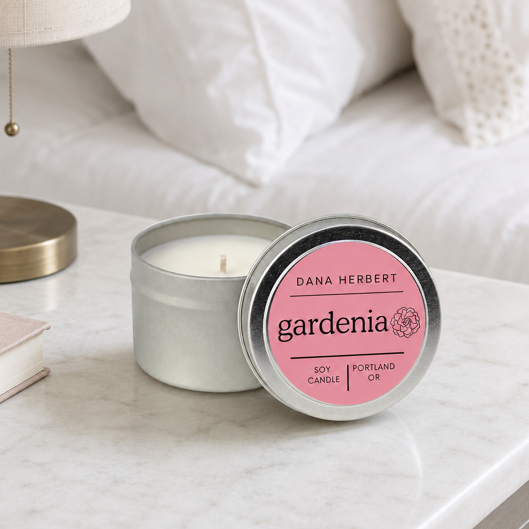A silver tin candle with its lid leaning beside it on a white marble surface. The lid has a pink label reading 'DANA HERBERT gardenia' and 'PORTLAND OR'.