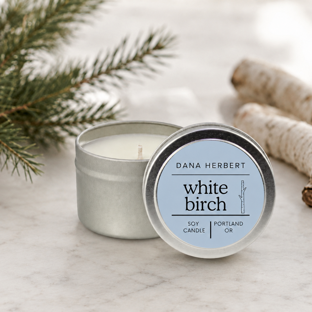 A silver metal tin candle with its lid placed beside it on a light marble surface. The lid label reads 'DANA HERBERT white birch' and 'SOY CANDLE | PORTLAND OR.